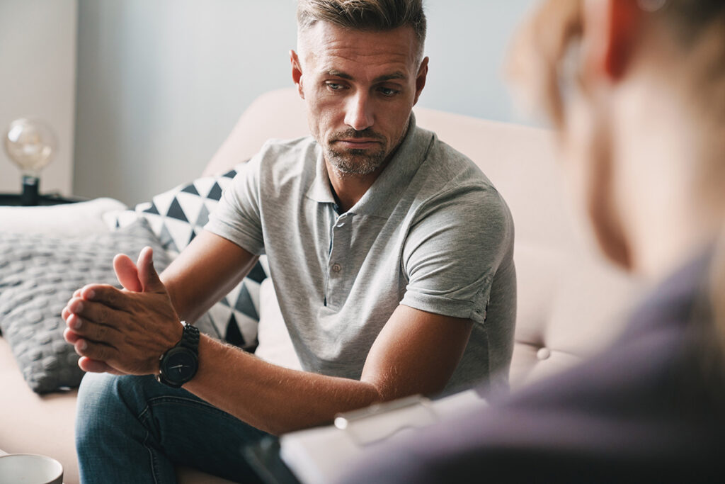 A man is sitting with his hands together against his knees in an individual therapy session.