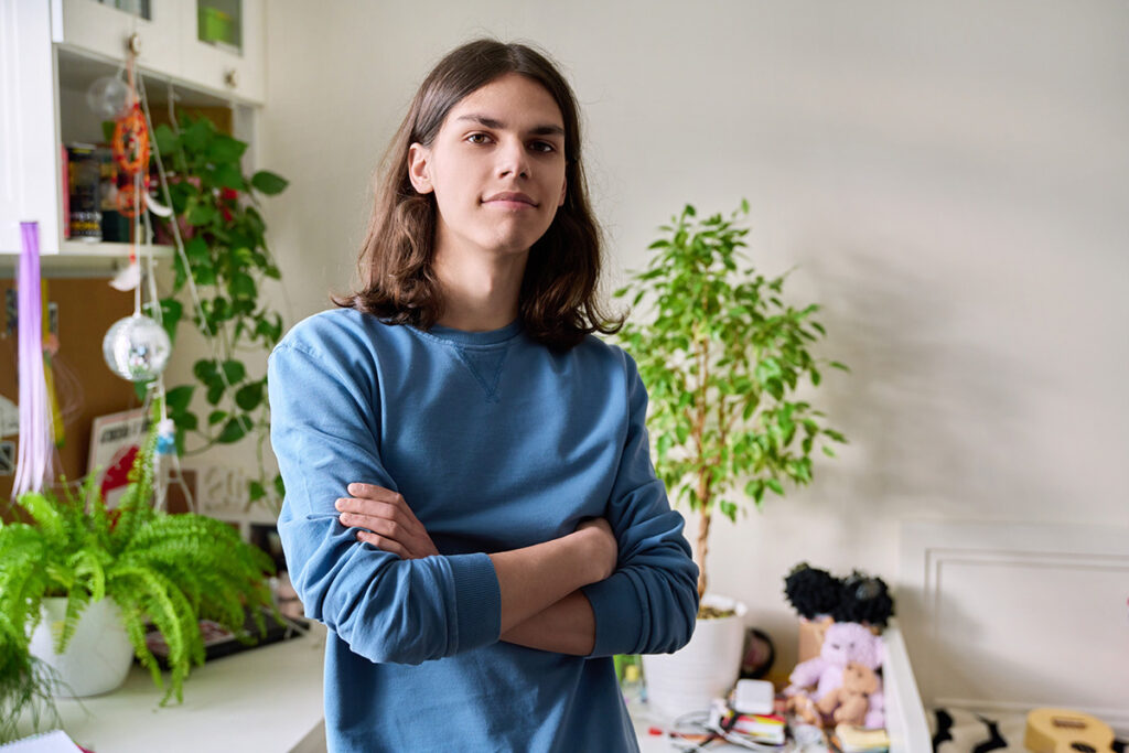 A teen with shoulder-length hair stands facing the camera with arms folded over their chest.