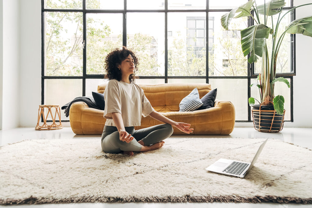 A woman is sitting cross-legged in a well-lit room on a white carpet, using a laptop to guide her through a meditation practice.