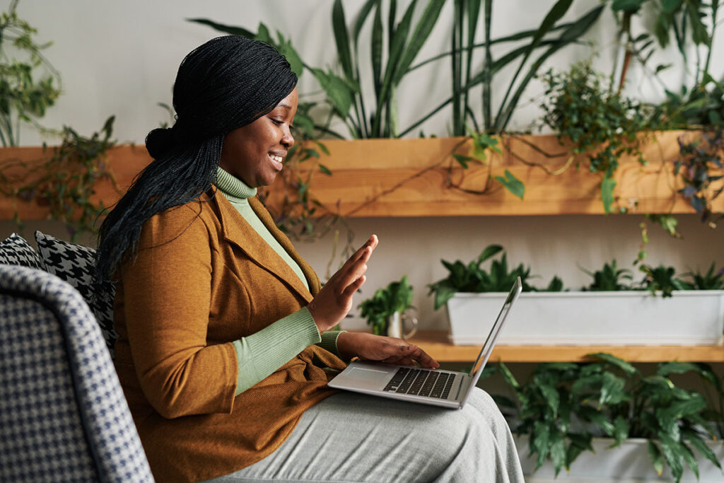 A woman is waving hello to a therapist she is speaking through her laptop. She is participating in alcohol addiction treatment.
