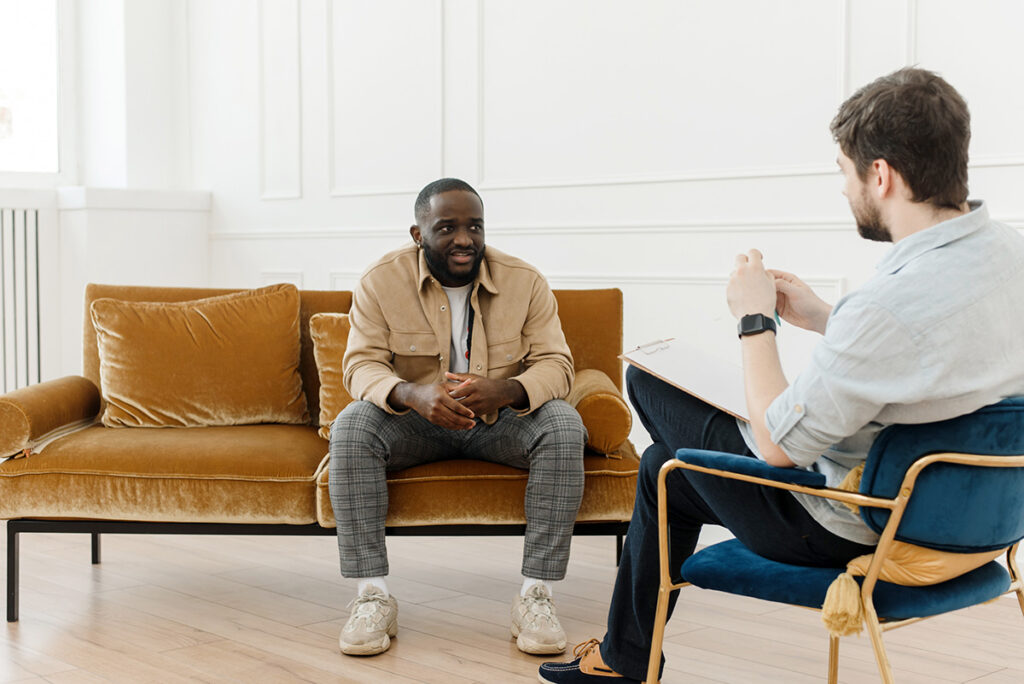A man is sitting on a bronze, velvet couch and speaking with a therapist sitting across from him.