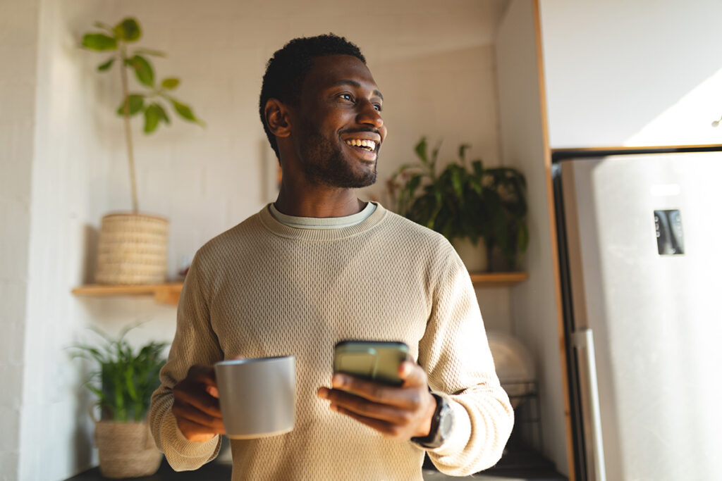 A man is smiling and holding a coffee mug in one hand and his phone in another.