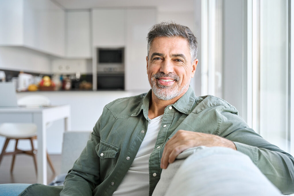 A man is sitting on his couch, just adjacent to his kitchen. He is smiling at ther camera, happy for the convenience of a intensive outpatient program