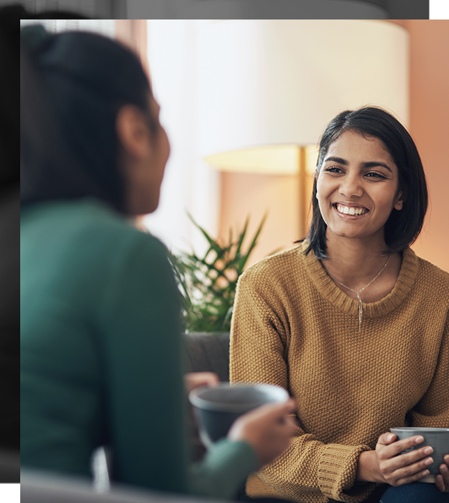 two women are sitting with mugs of coffee and smiling.
